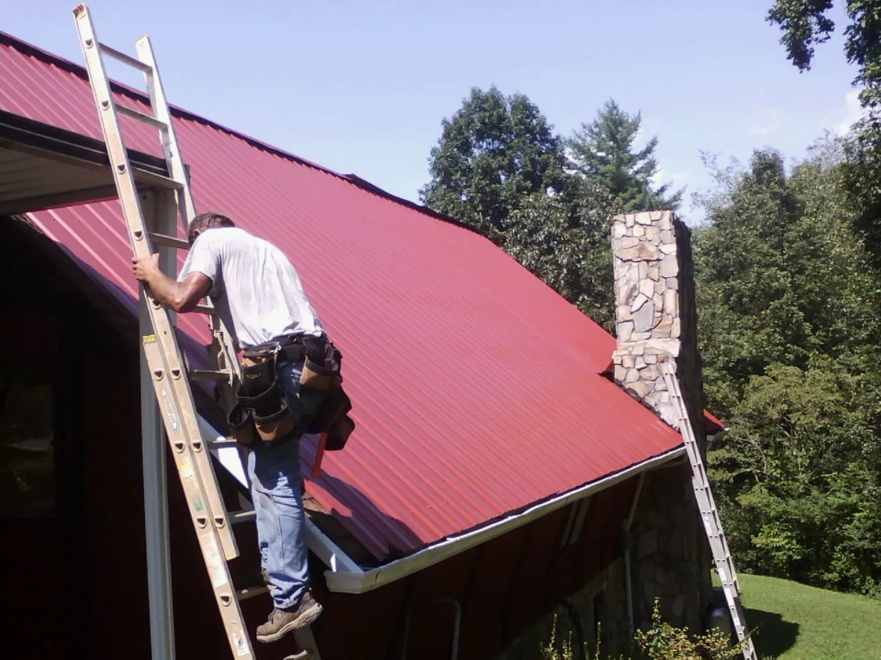 Roofer on a ladder beside a red metal roof and stone chimney — Soffit & Fascia Repair by Roofing Brothers in New Ulm